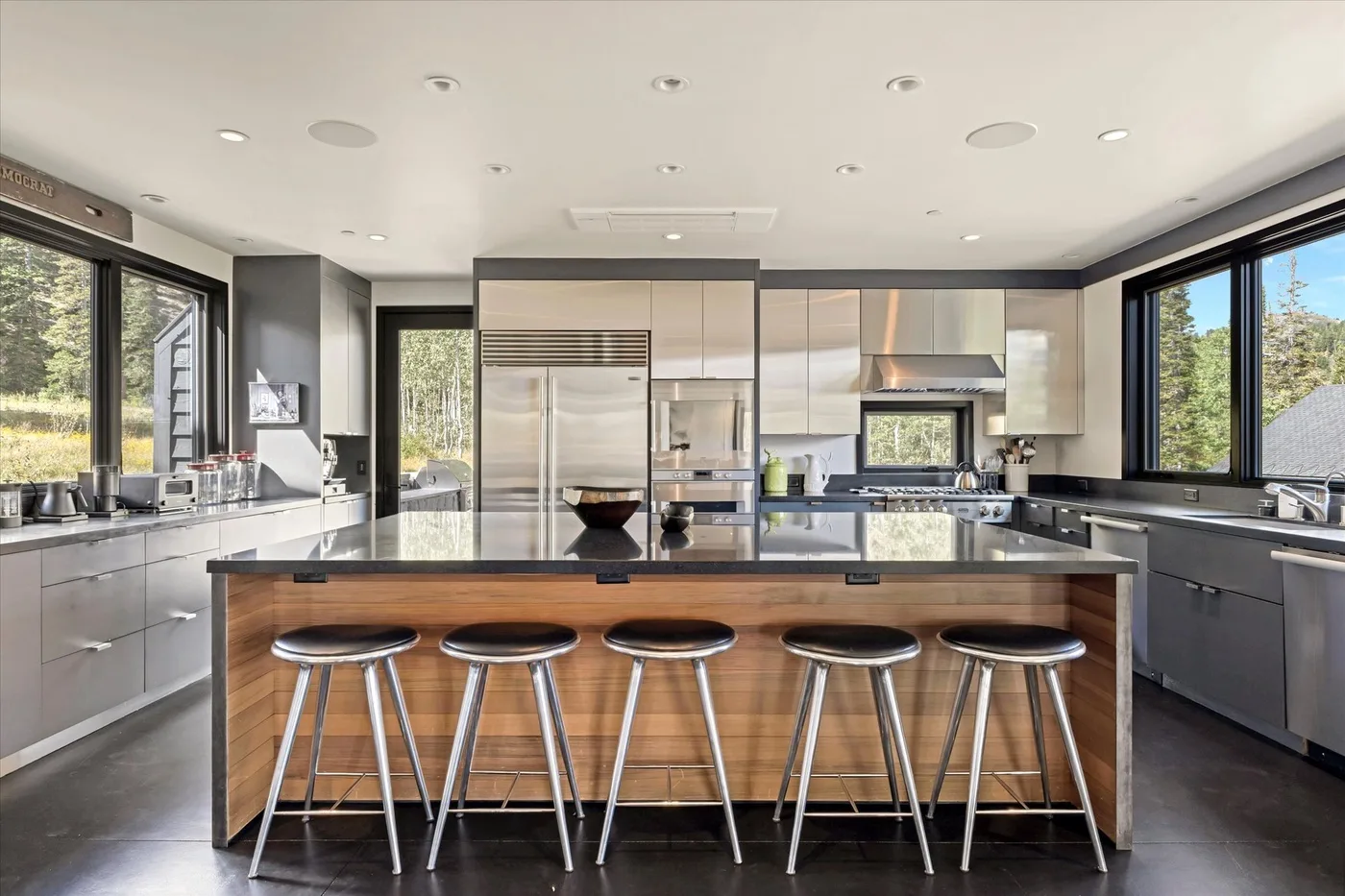 A modern kitchen with a long island and metal stools sits between large windows.