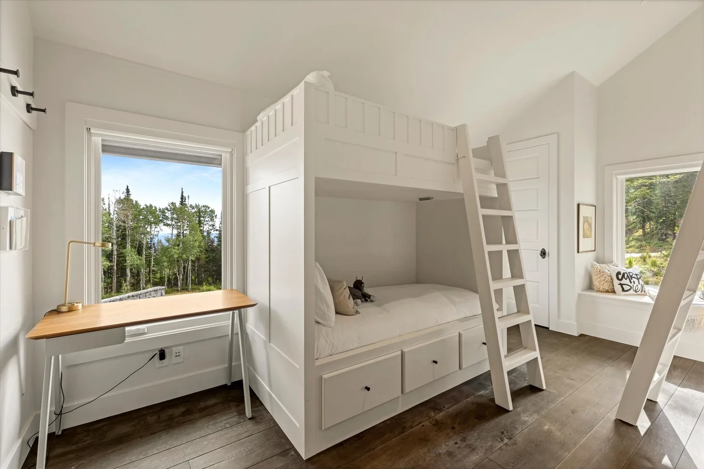 A bright bedroom features a white bunk bed and a desk beside the window.