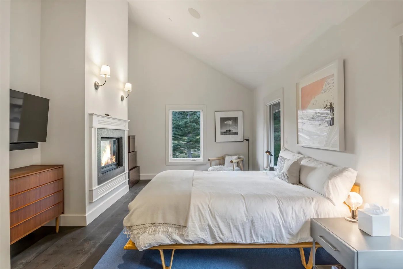 A bright bedroom with a large bed, a fireplace, and windows overlooking greenery.
