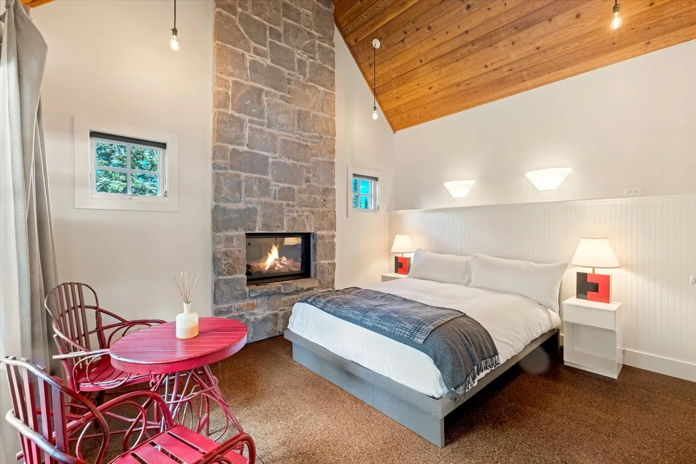 A bedroom with a bed, stone fireplace, and red seating beneath a wood ceiling.