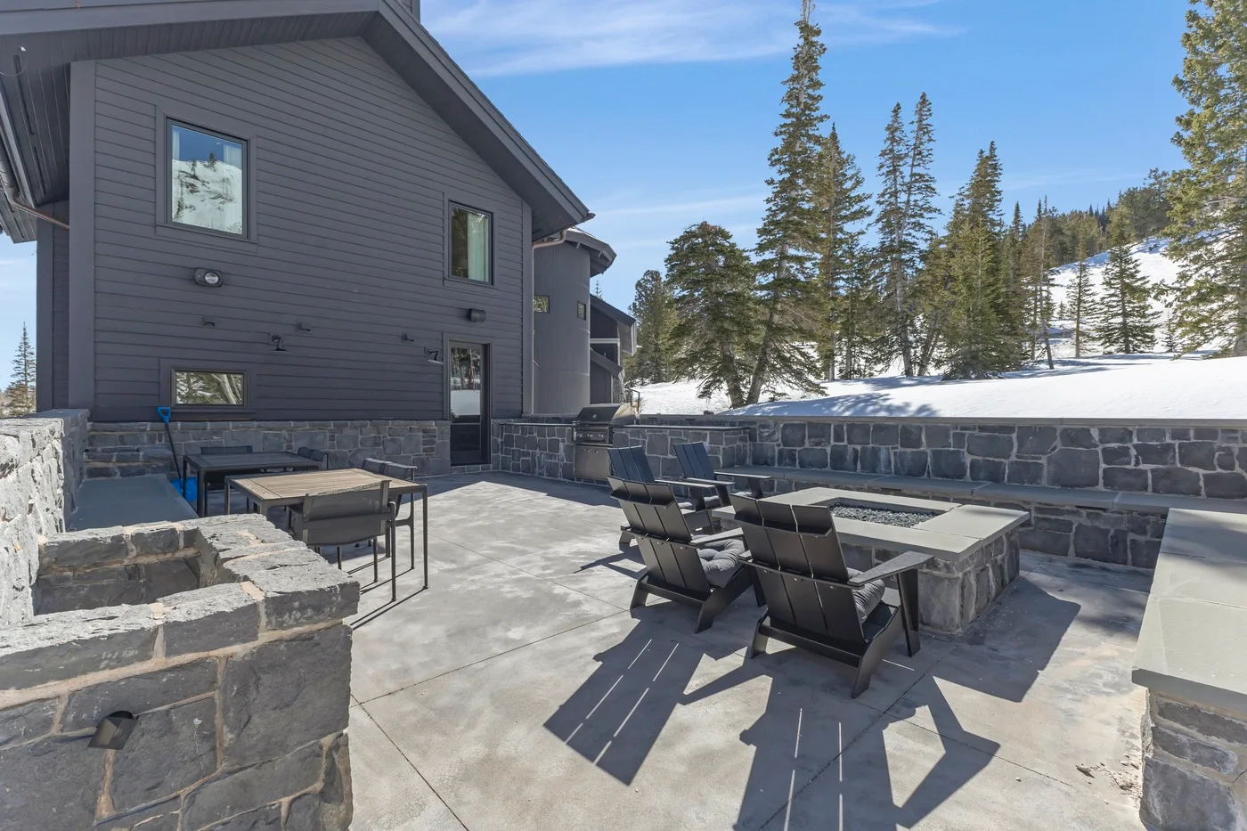 An outdoor stone patio with seating beside a dark-sided building and snowy trees.