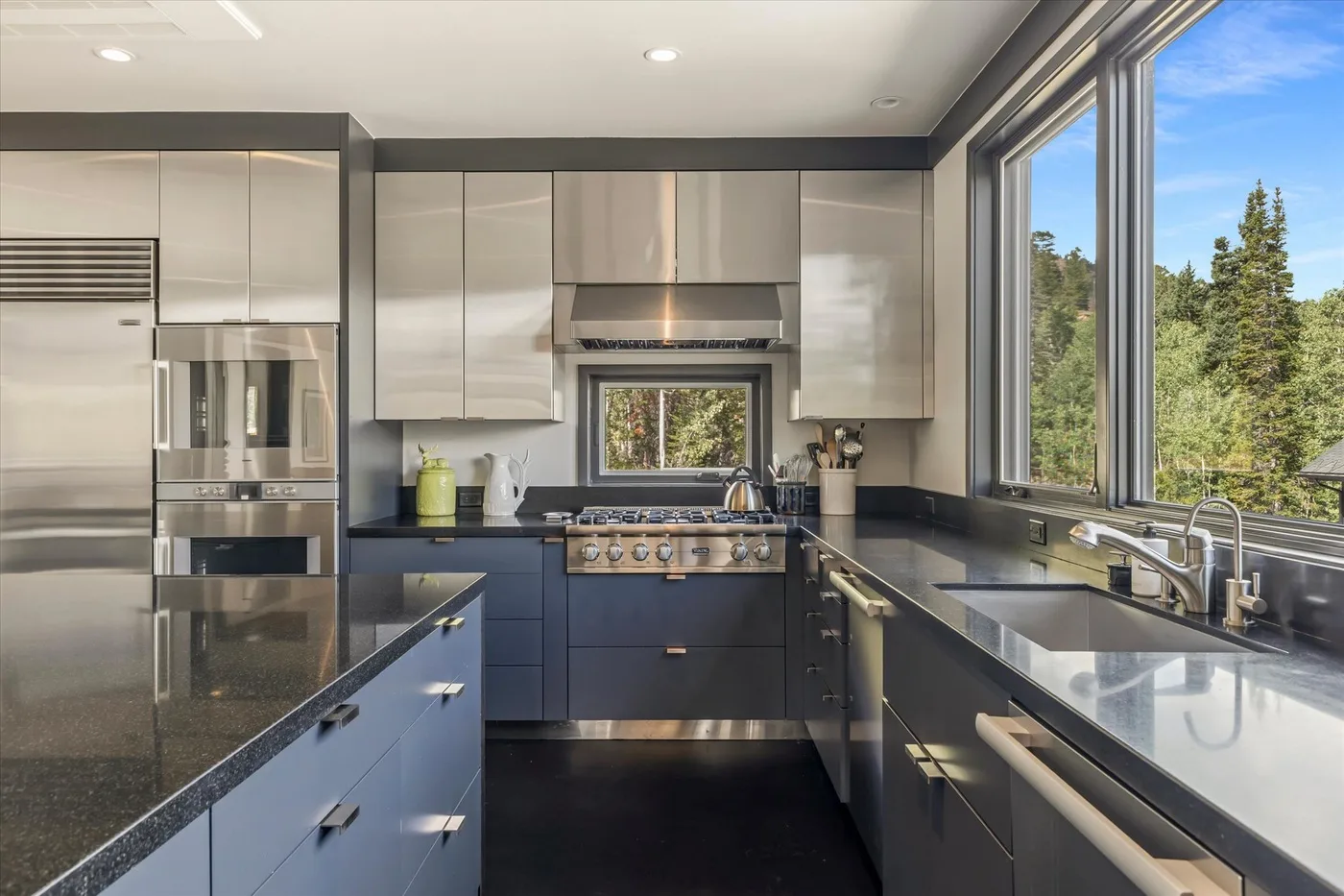 A modern kitchen with dark counters and large windows overlooking trees.