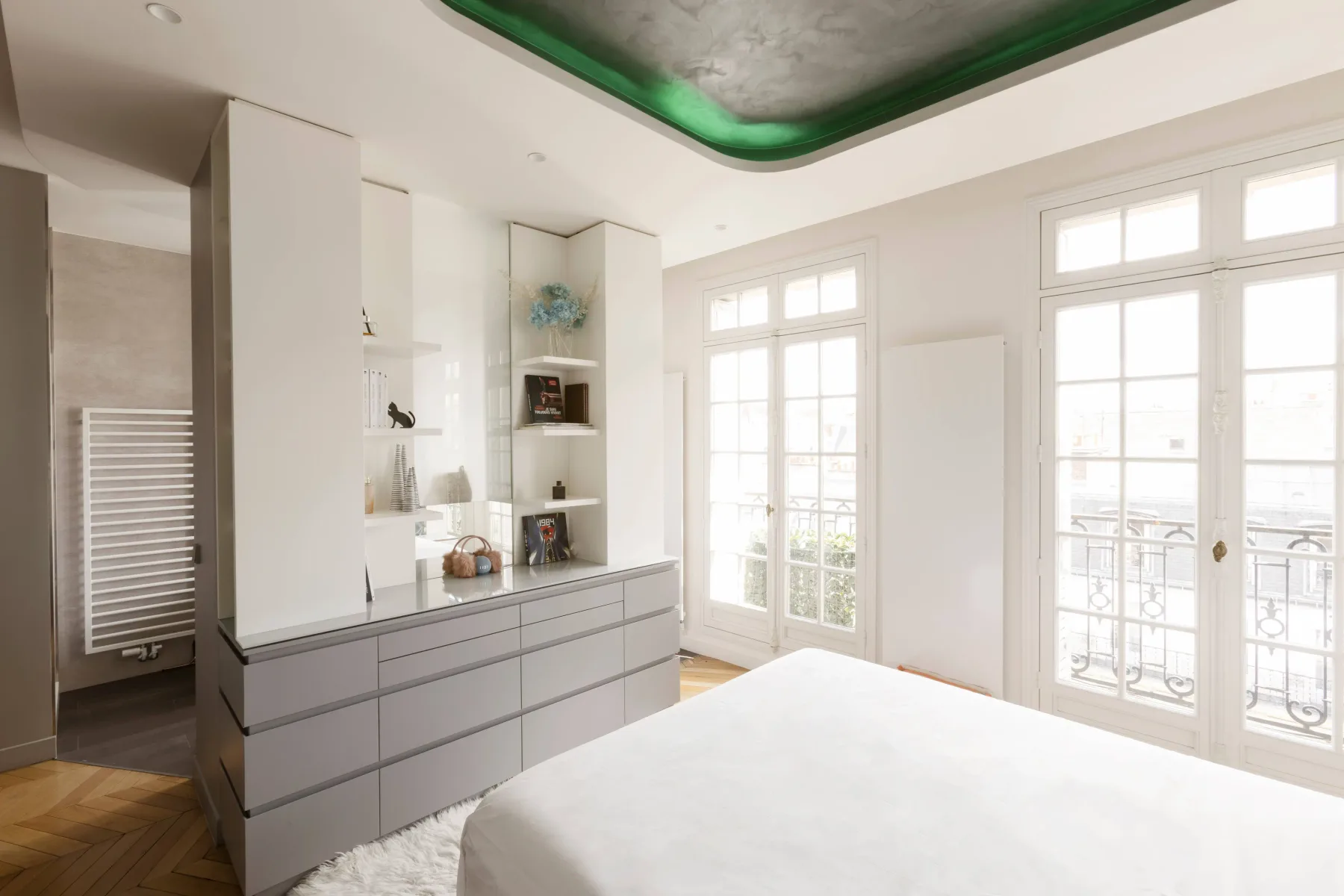 A bright bedroom with a bed, built-in shelving, and tall windows.