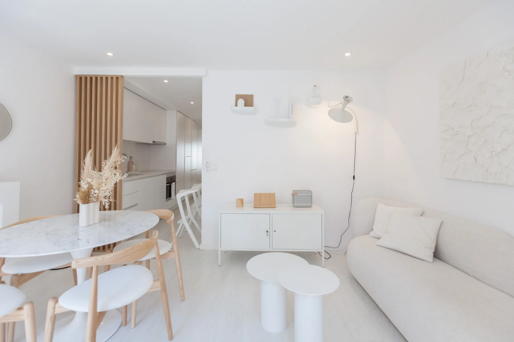 Una luminosa sala de estar y comedor con un sofá claro, mesas redondas y una cocina abierta.