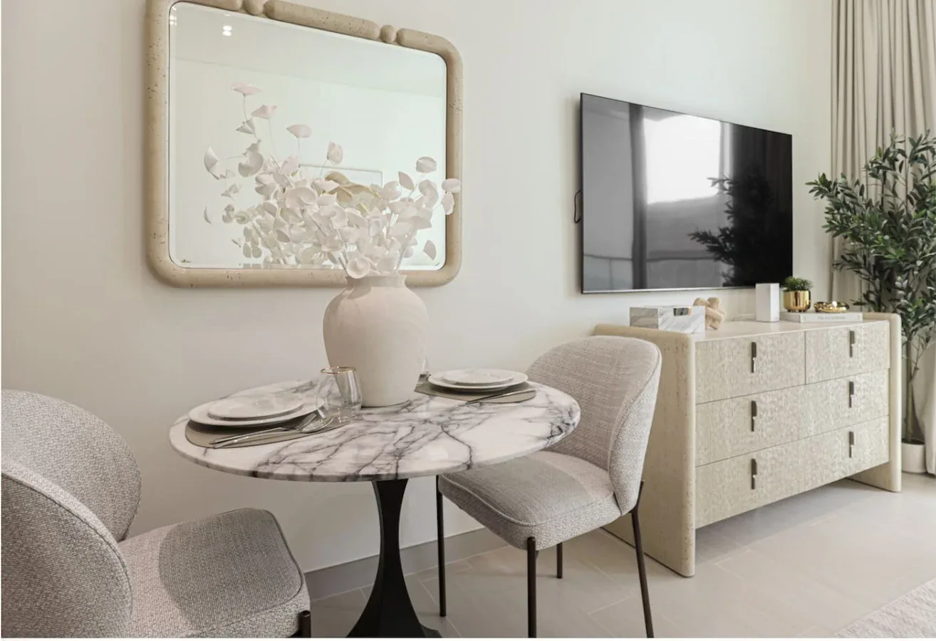 A bright dining area with a round table, upholstered seating, and a television.