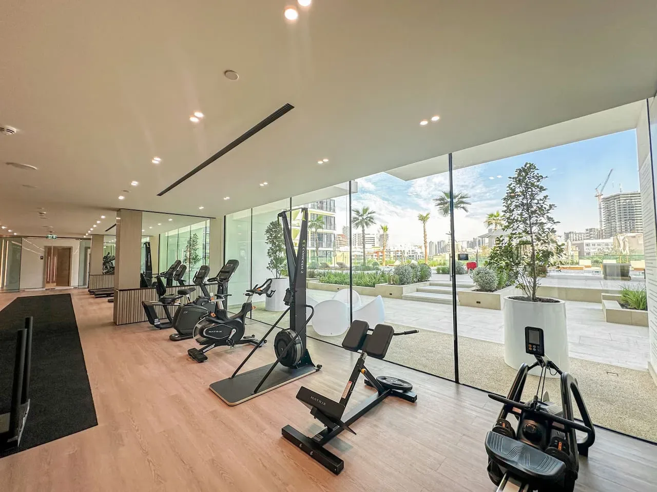 A bright fitness room with exercise equipment beside floor-to-ceiling windows.