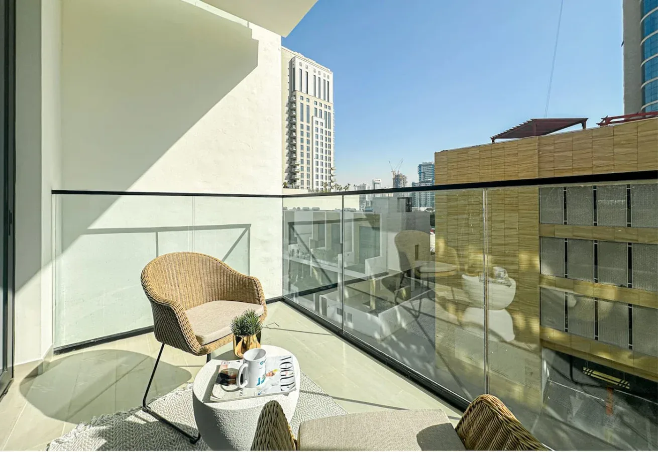 A sunlit balcony with outdoor seating and a glass railing overlooks nearby buildings.