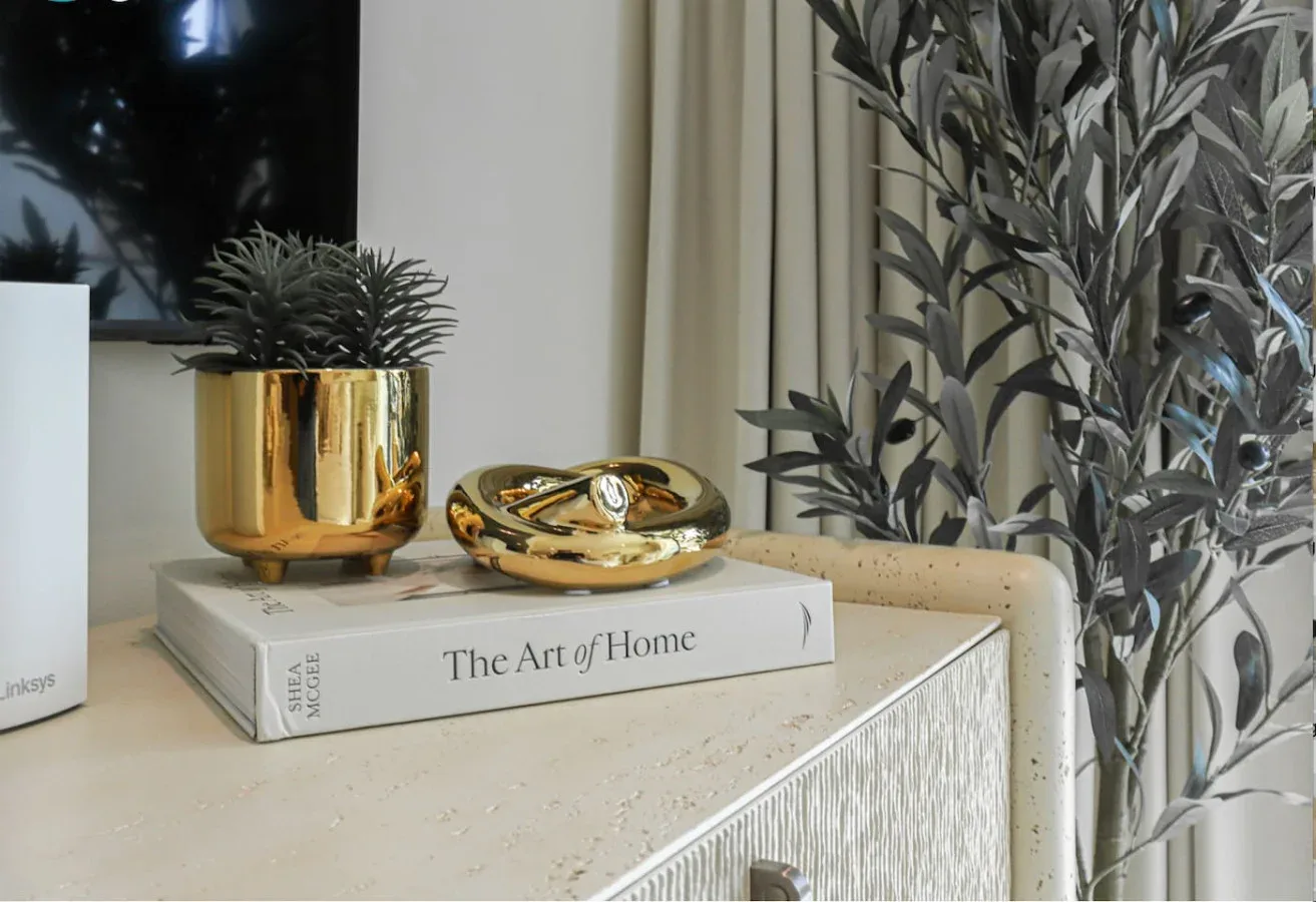 A living room console displays a gold planter and decorative objects beside leafy foliage.