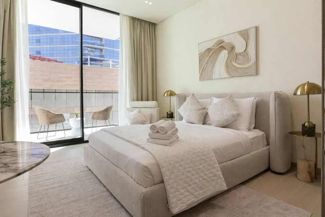 A bright bedroom features a neatly made bed beside large windows and a small balcony.