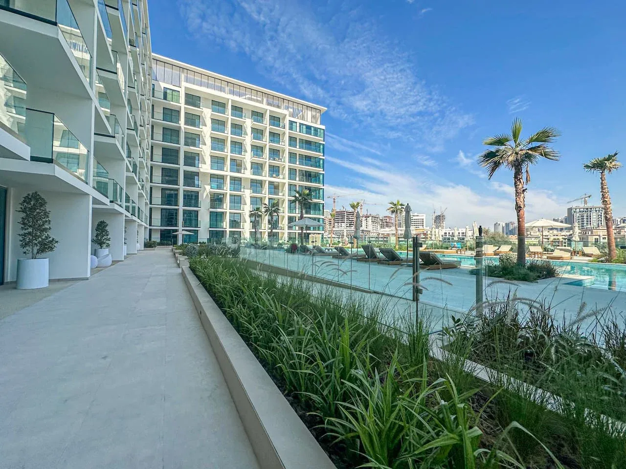 A modern building and outdoor pool area beside a paved walkway and landscaped greenery.