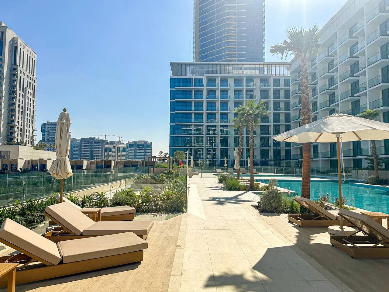 A sunny outdoor pool terrace with lounge seating, umbrellas, and modern buildings.