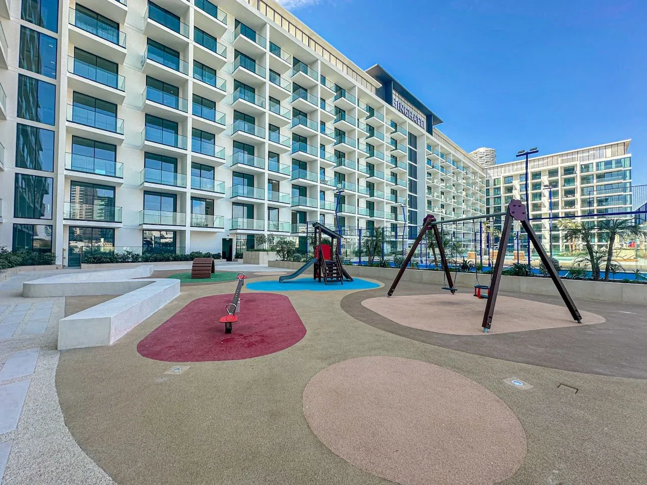 A playground with swings and play equipment sits in front of a modern apartment building.