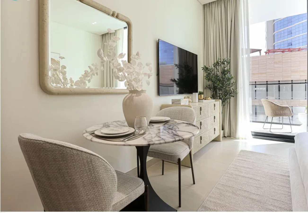 A bright dining and living area with a round table, television, and large windows.