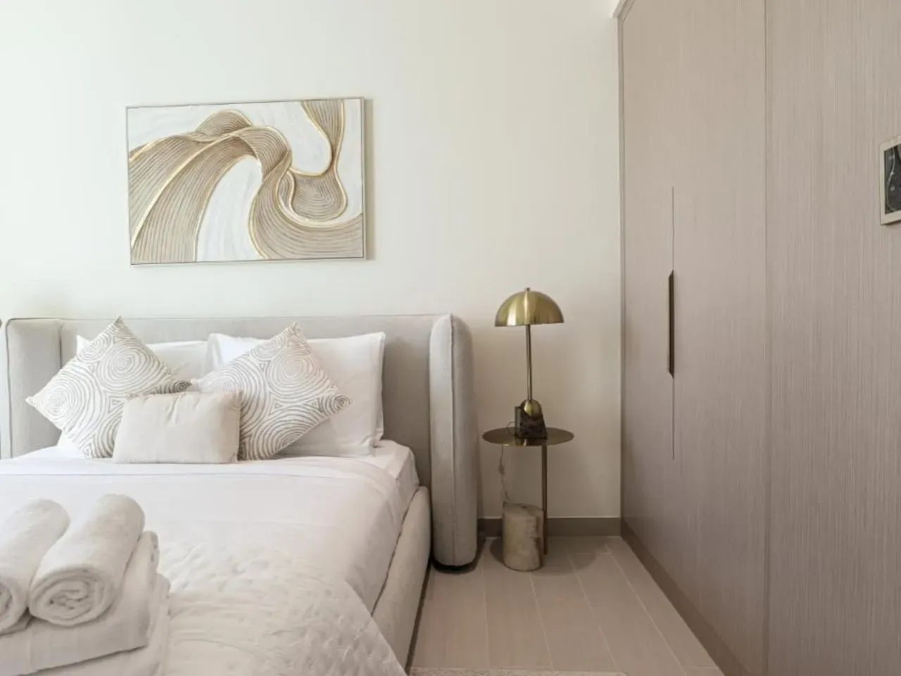 A bright bedroom with a neatly made bed, decorative pillows, and abstract wall art.