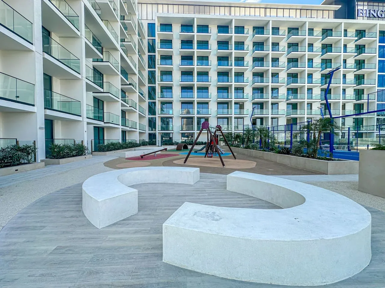 A courtyard playground sits among modern buildings, with curved white seating in the foreground.