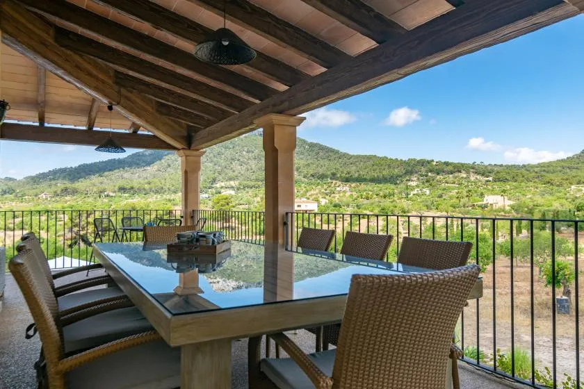 Una zona de comedor exterior cubierta, con mesa y asientos, tiene vistas a una colina verde.