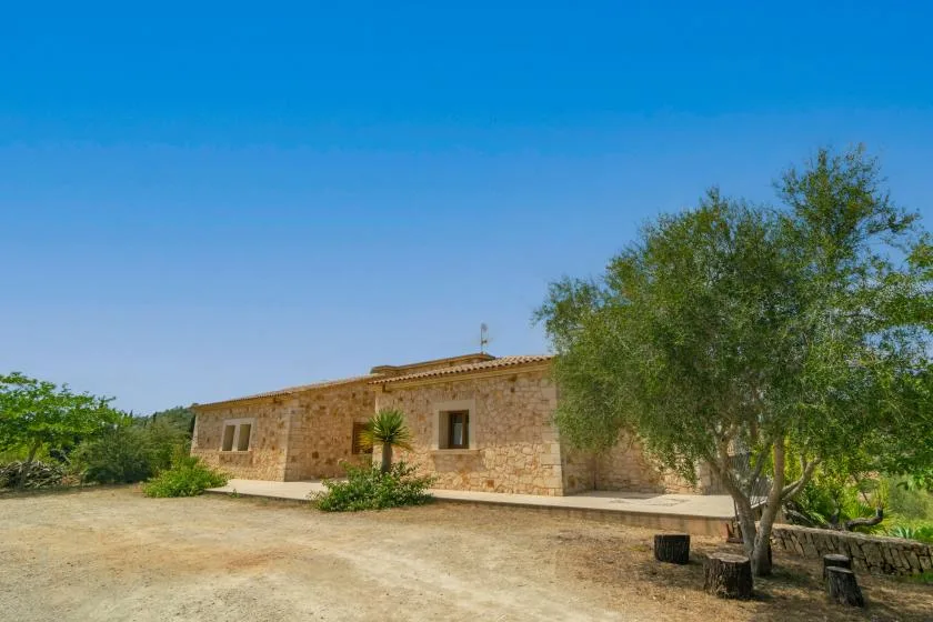 Una casa de piedra está junto a un patio seco y árboles frondosos.