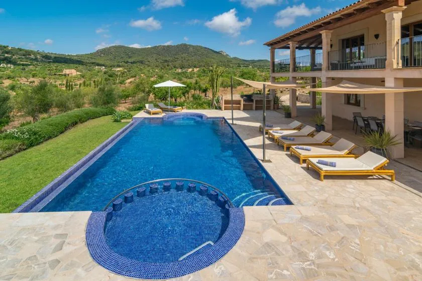 Una piscina exterior y un spa circular están junto a una terraza con vistas a colinas verdes.