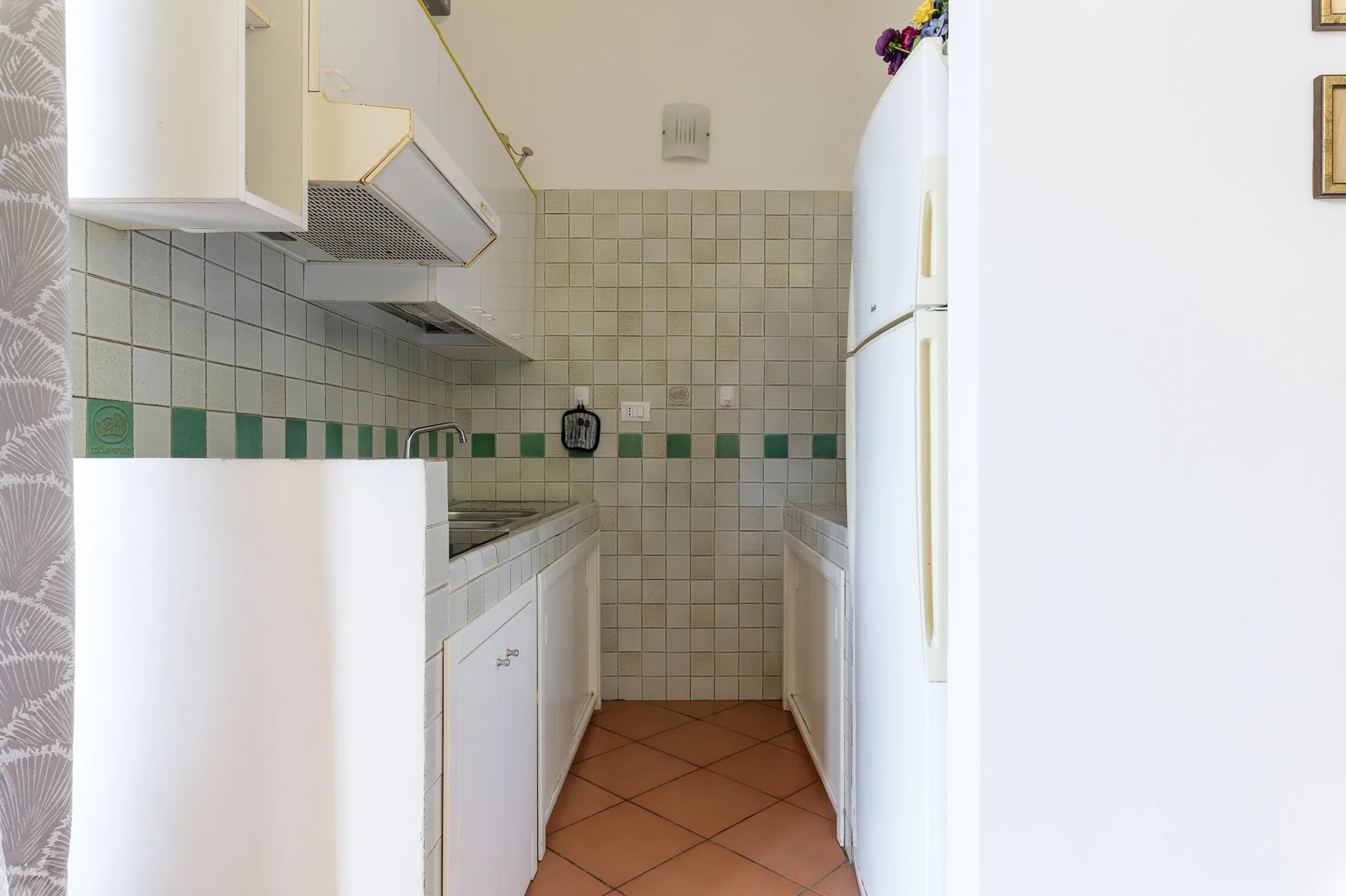 A narrow tiled kitchen with counters and a refrigerator.