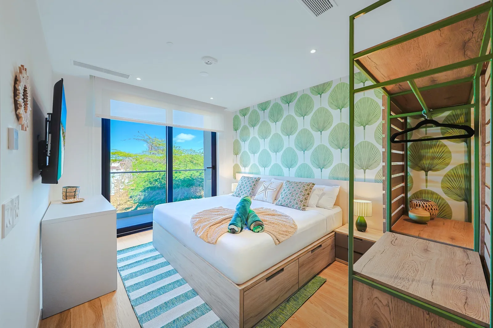 Unwind in your coastal-inspired bedroom with bright tropical wallpaper, a cozy bed adorned with playful towel art, and lush garden views framing your private retreat.
