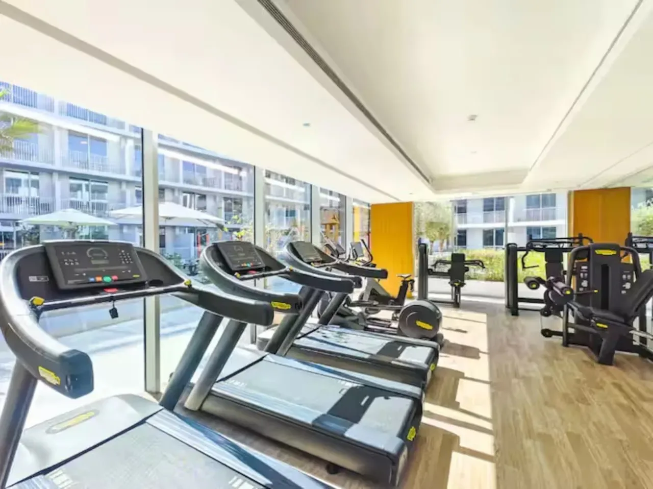 Un gimnasio luminoso con cintas de correr y otros equipos de ejercicio junto a grandes ventanales.