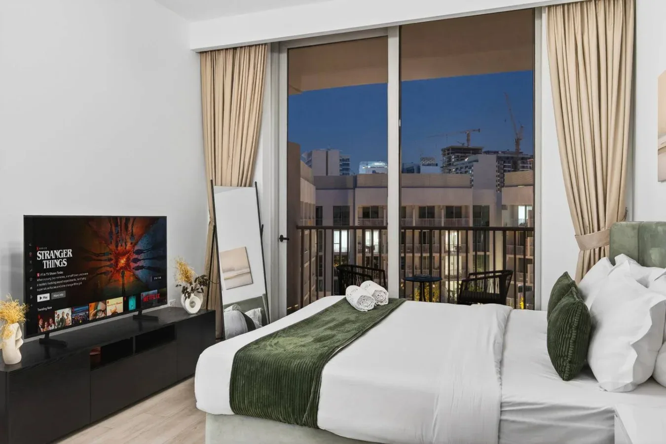 Dormitorio con una cama bien tendida y grandes ventanas con vistas a edificios cercanos.
