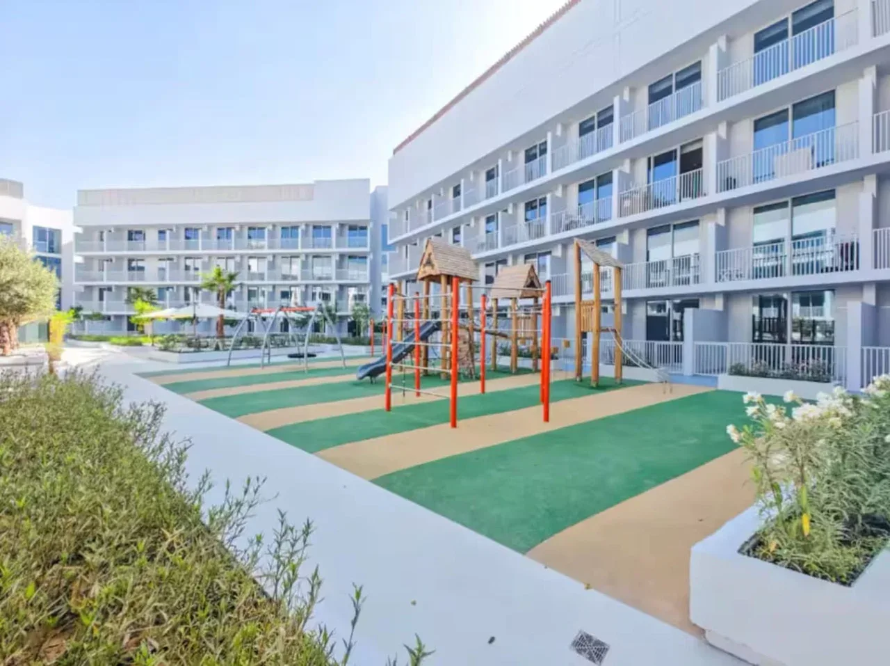 Un parque infantil al aire libre con estructuras de colores ocupa un patio ajardinado rodeado de edificios de apartamentos.