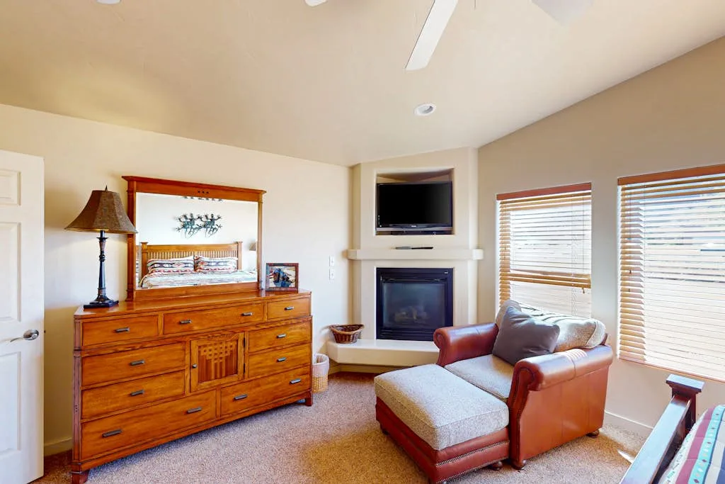 A bright living room with a fireplace, wooden dresser, and seating beside large windows.