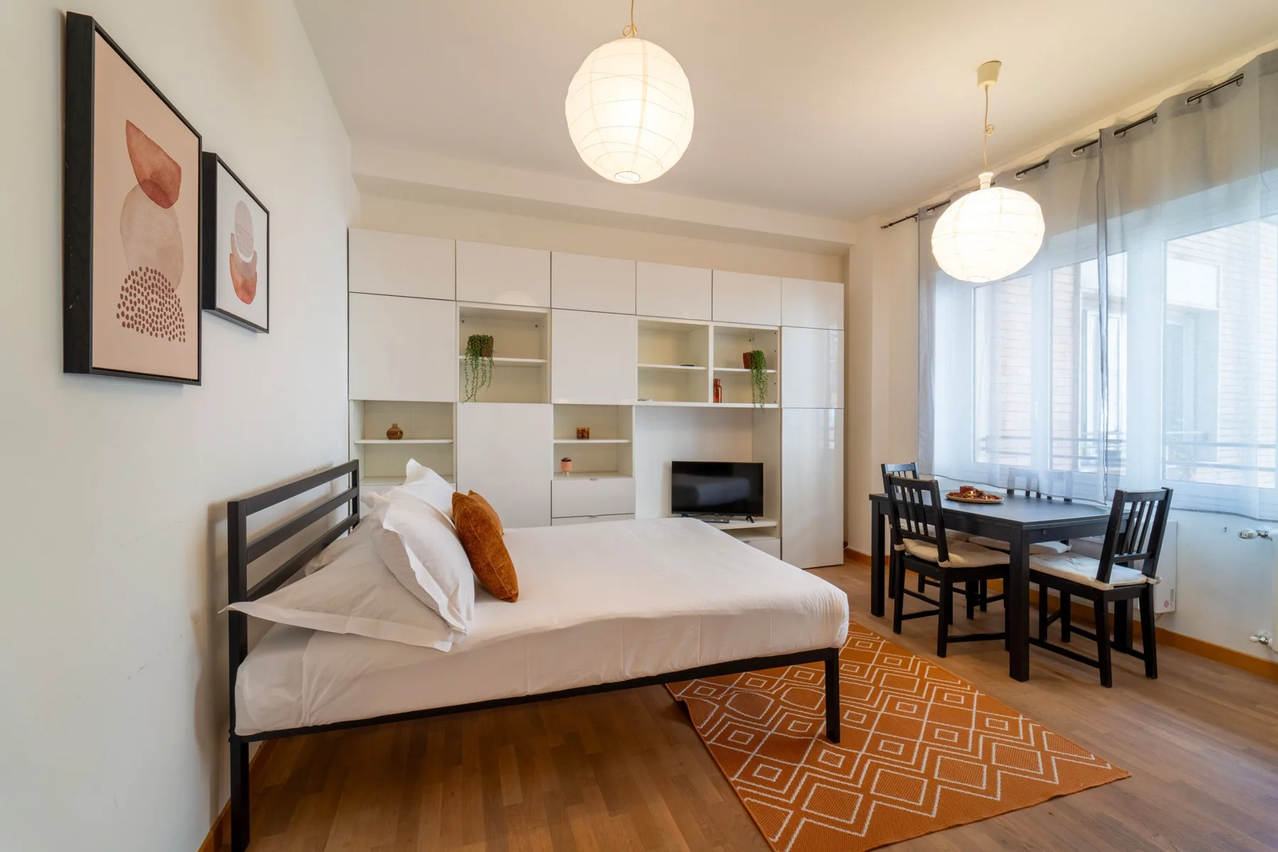 Un dormitorio luminoso con cama, estanterías empotradas y una mesa junto a las ventanas.