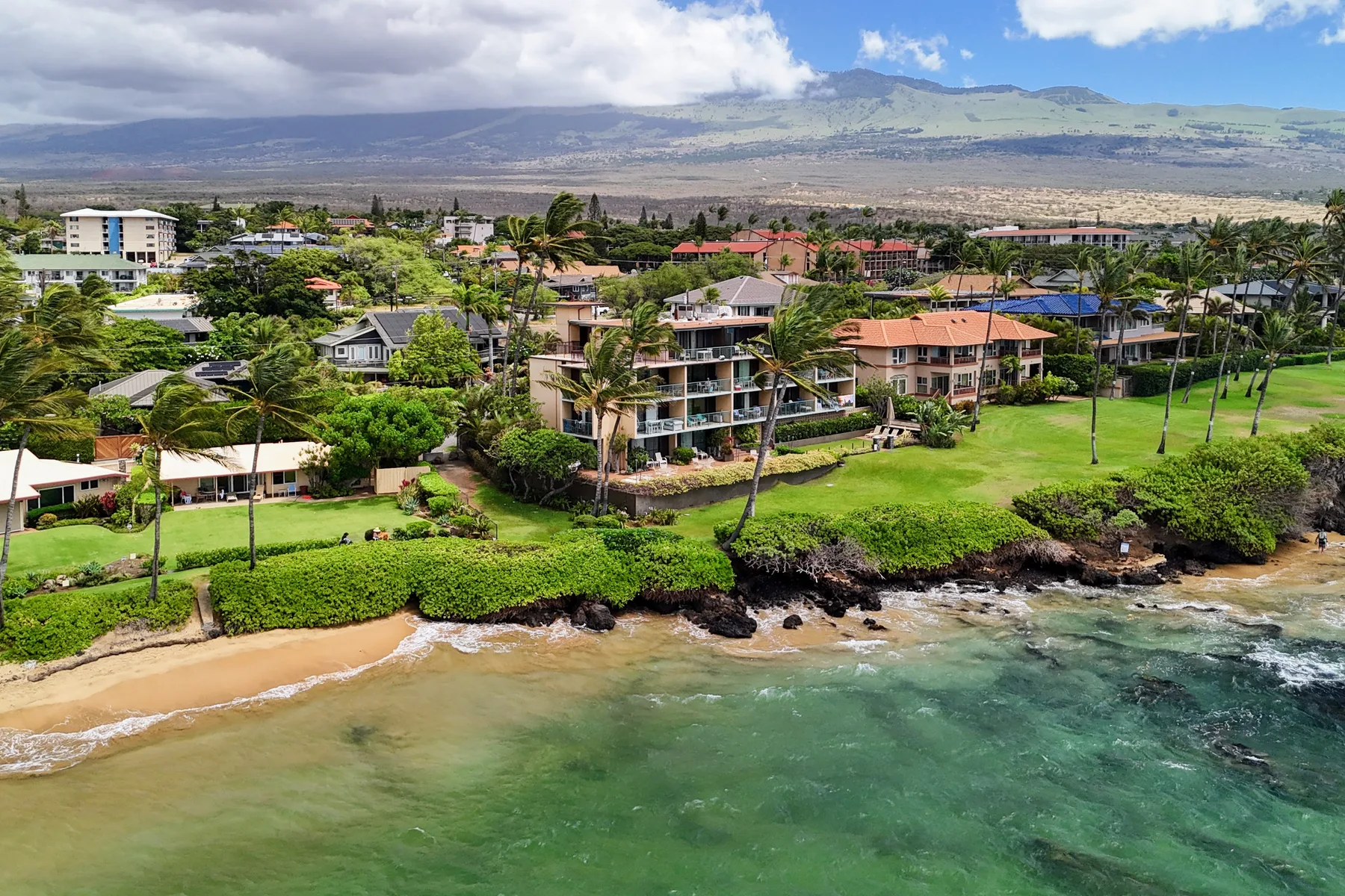 A coastal neighborhood with beachfront buildings, green lawns, and mountains in the distance.