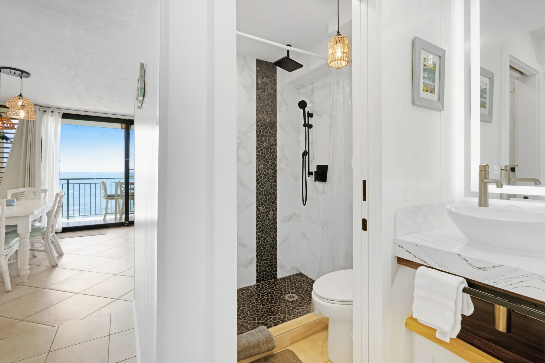 A bright bathroom with a glass shower, toilet, and vanity beside a dining area.