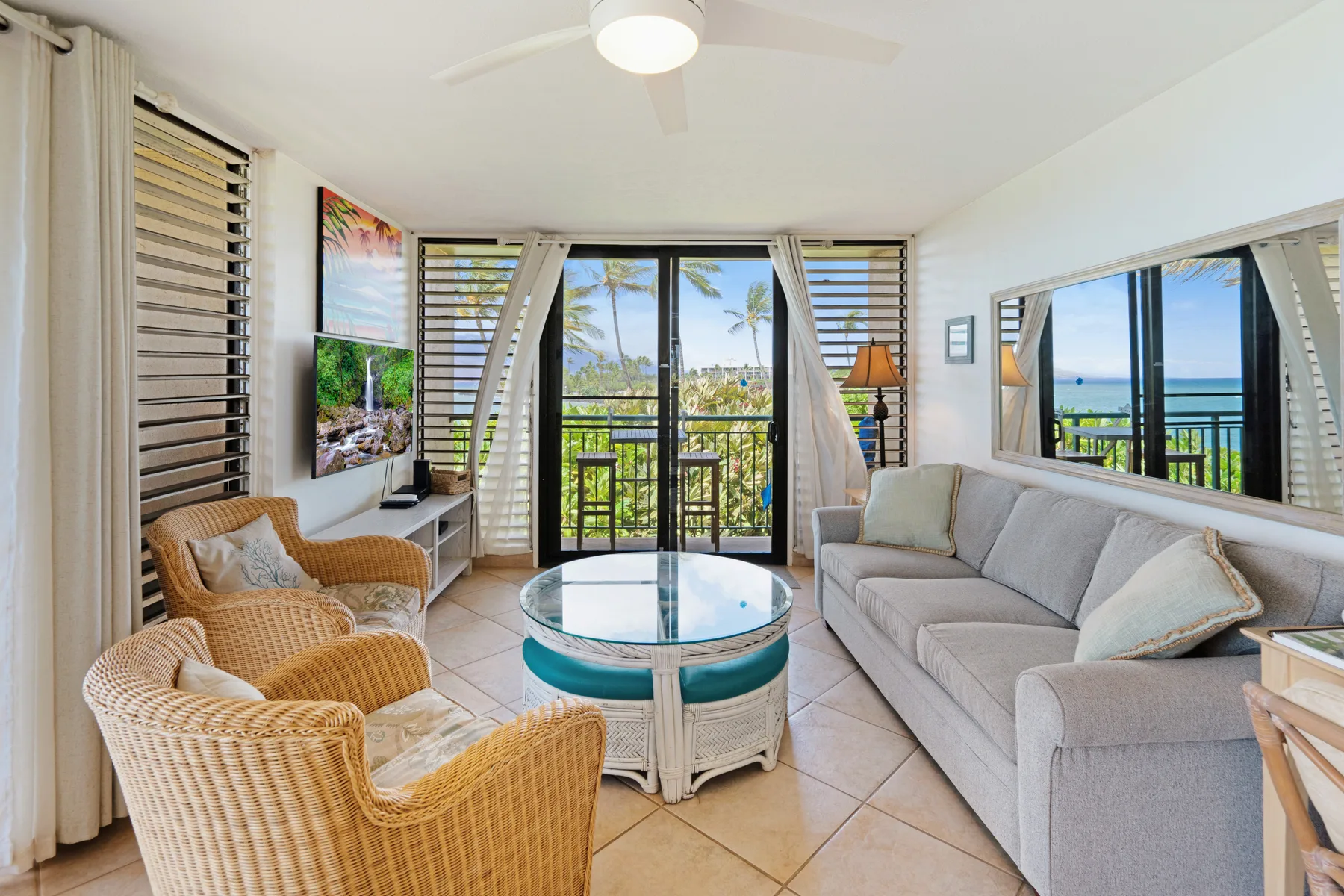 A bright living room has seating around a glass coffee table, with windows overlooking the water.