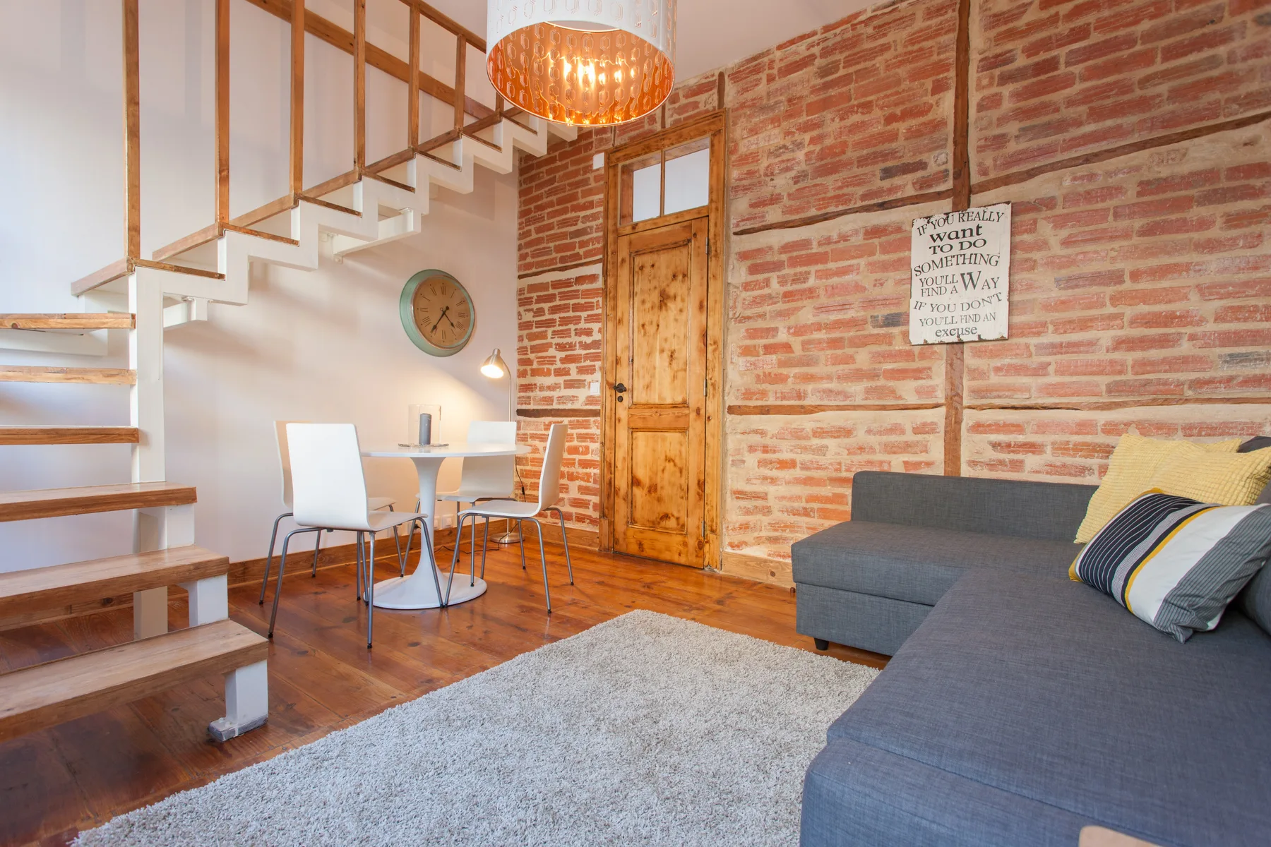 A living and dining area with a gray sectional sofa beside an exposed brick wall.