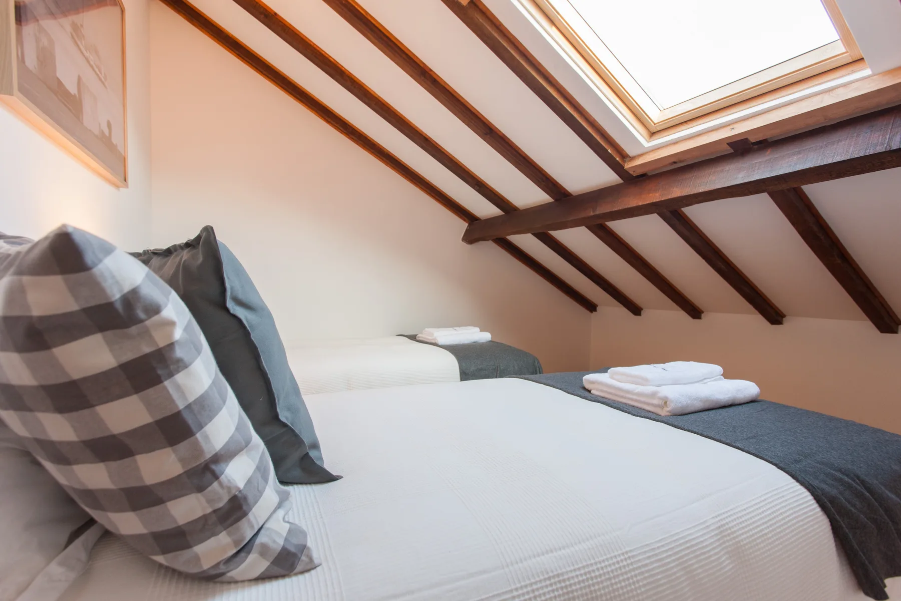 A bedroom with a bed beneath a sloped, beam-lined ceiling and skylight.