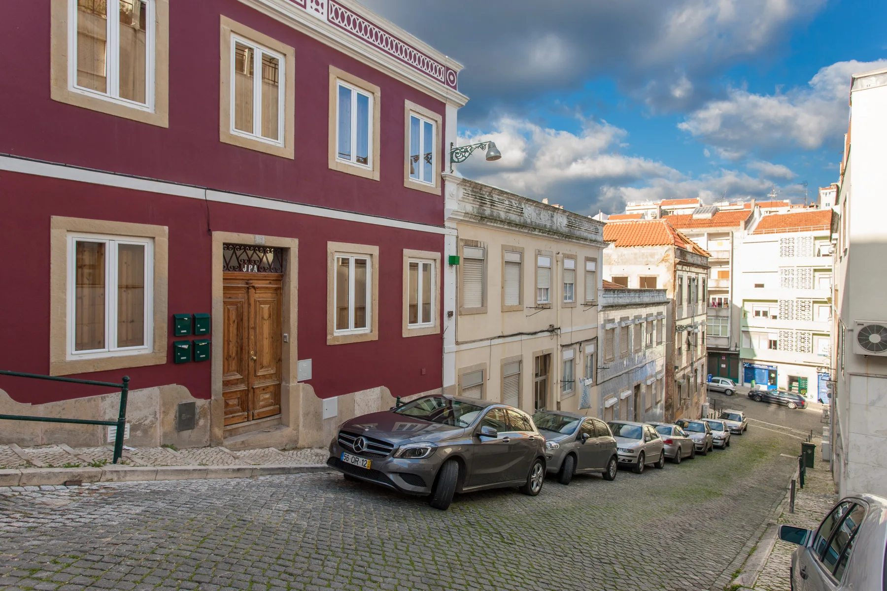 A cobblestone street slopes between colorful urban buildings, with parked cars along the curb.