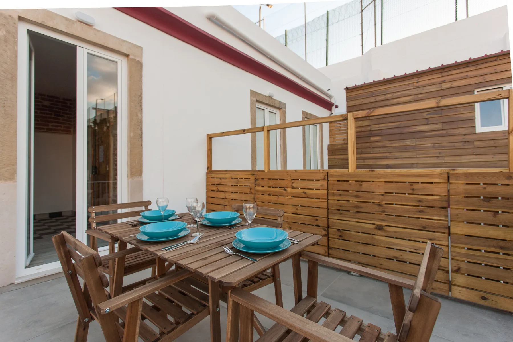 A wooden dining table and seating sit on a paved terrace beside white walls.
