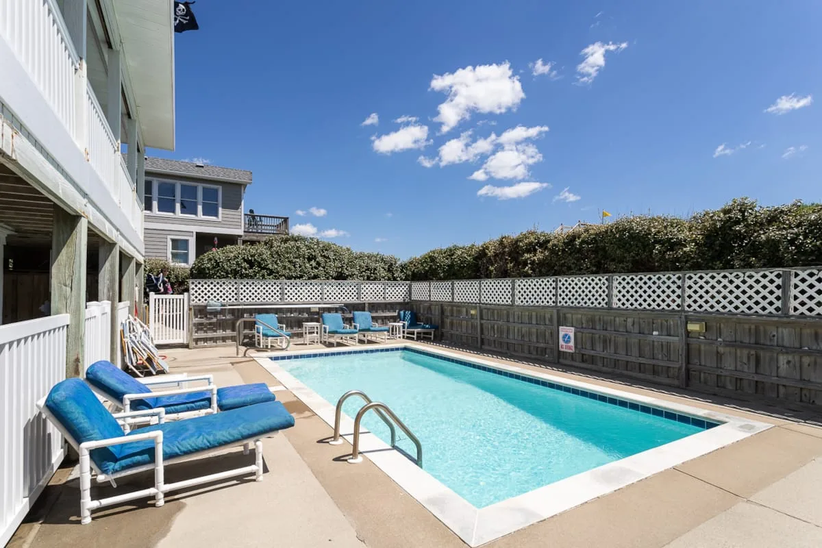 An outdoor swimming pool is surrounded by lounge seating and a tall fence.