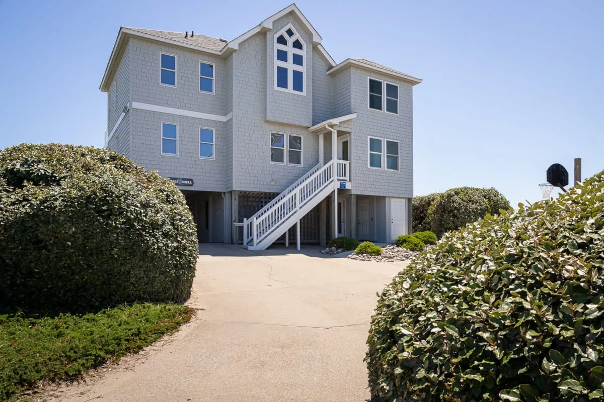 A raised coastal-style house stands behind a paved driveway and shrubs.