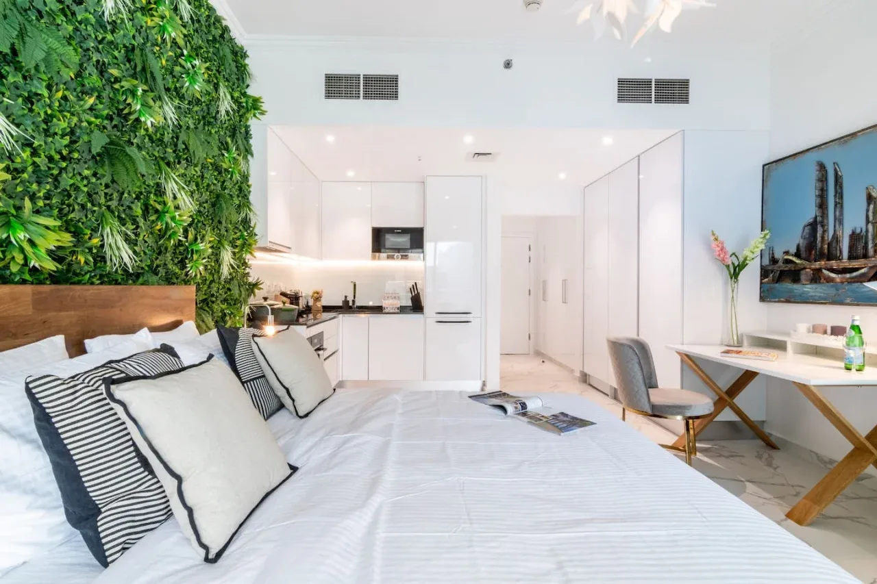 A bright bedroom with a large bed, leafy wall, and kitchenette in the background.