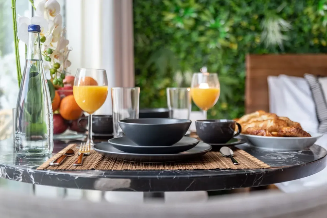 A breakfast table with bowls, fruit, juice, and pastries beside a leafy backdrop.