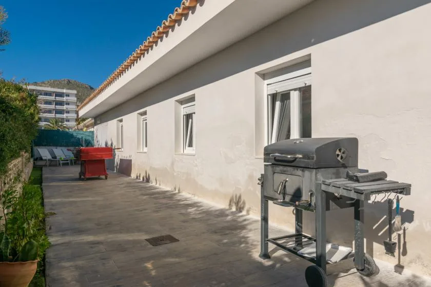 A paved outdoor patio runs alongside a white building, with a barbecue grill nearby.