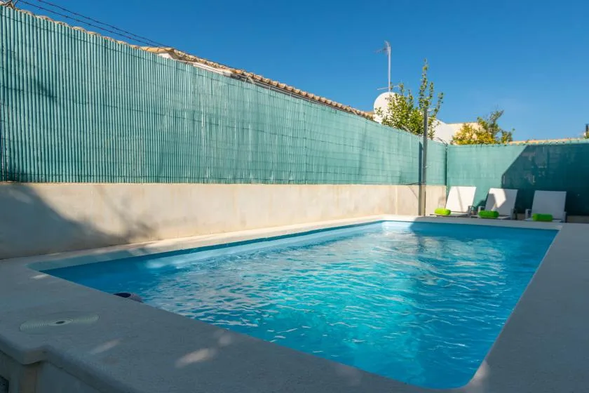 An outdoor swimming pool sits beside a tall privacy fence under a clear sky.