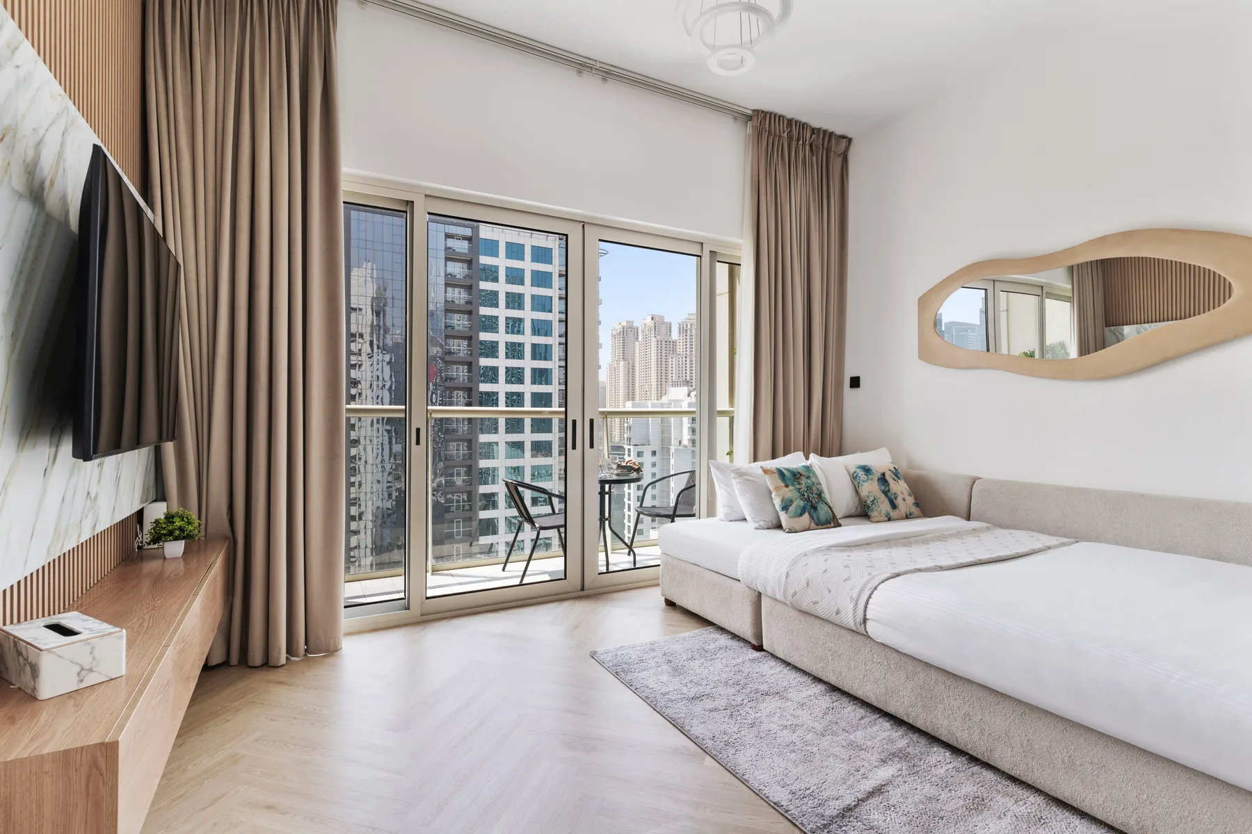 Un dormitorio luminoso con una cama, un televisor y grandes ventanas con vista a edificios cercanos.