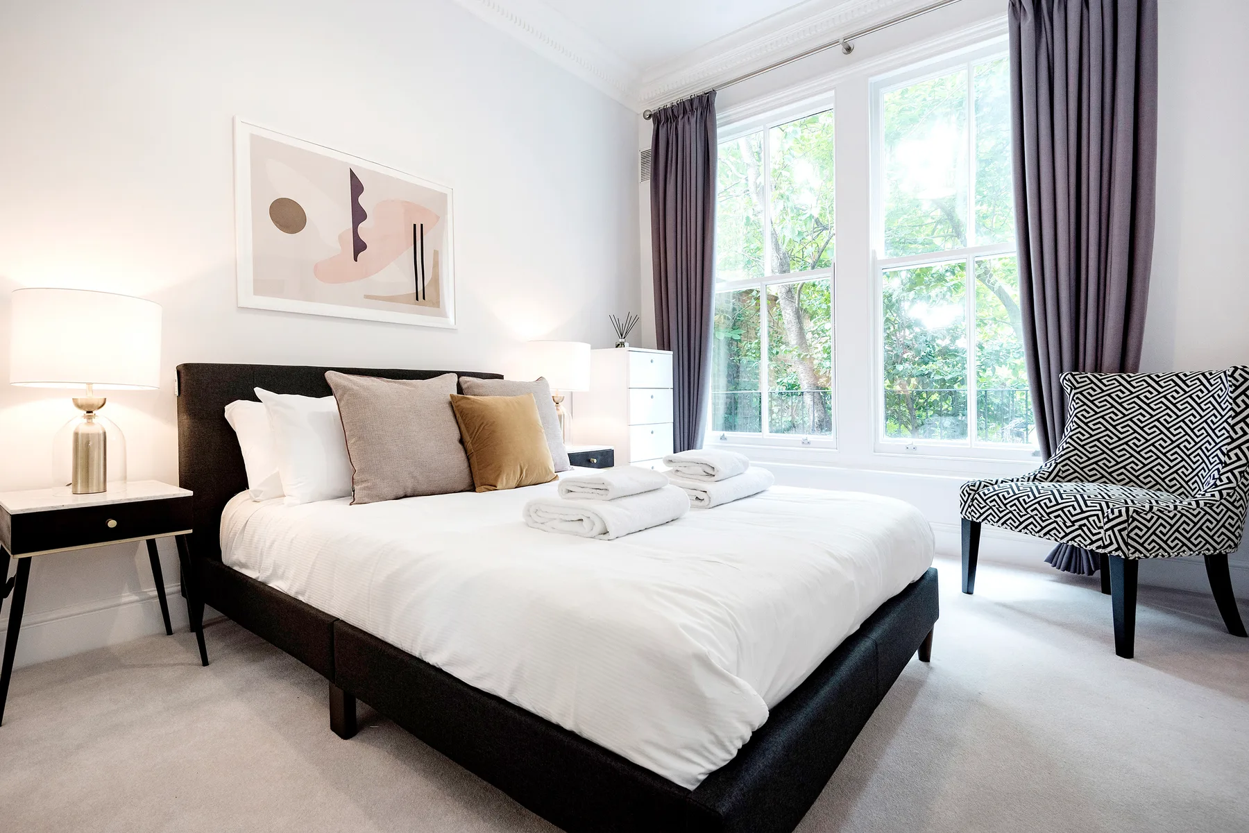 Un dormitorio luminoso con una cama bien tendida junto a grandes ventanas.