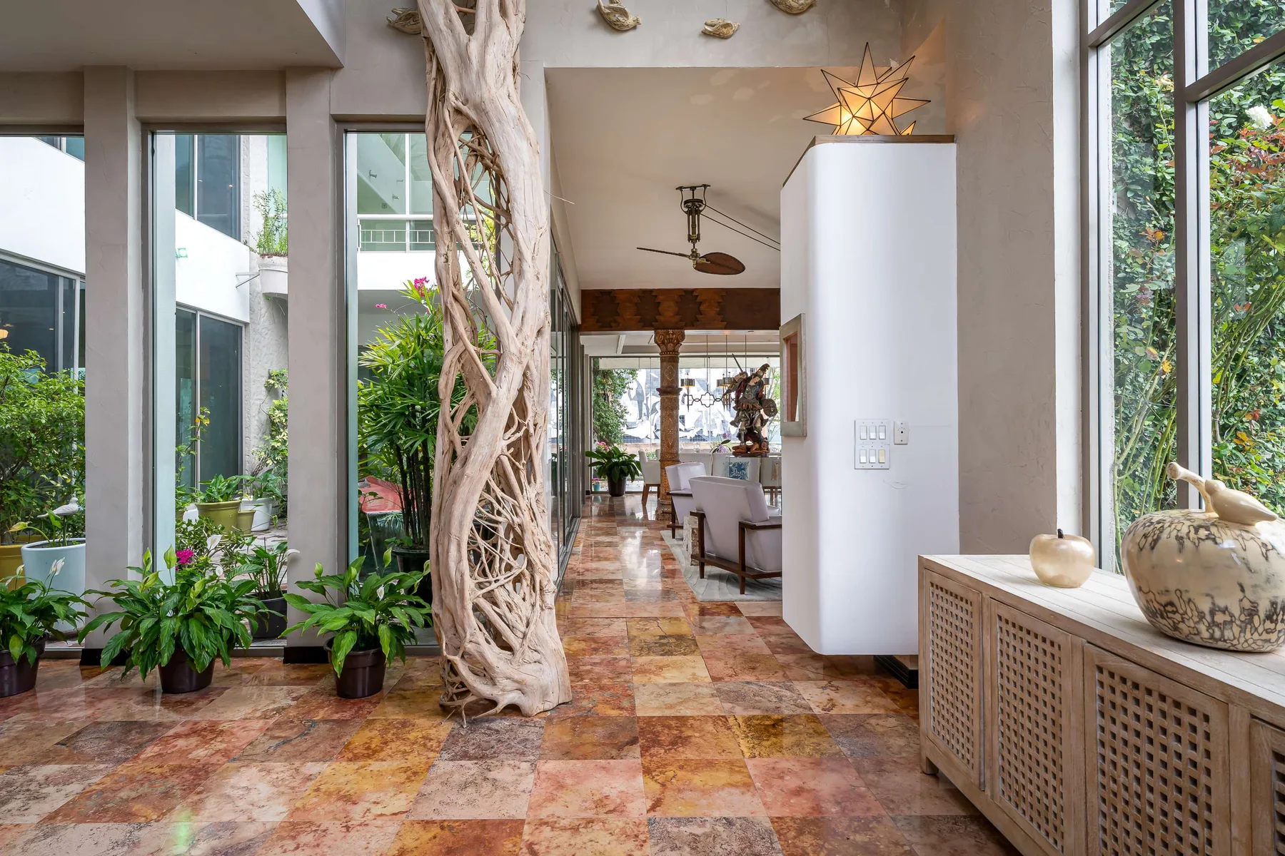 Un pasillo interior luminoso con grandes ventanas, plantas en macetas y un árbol central.