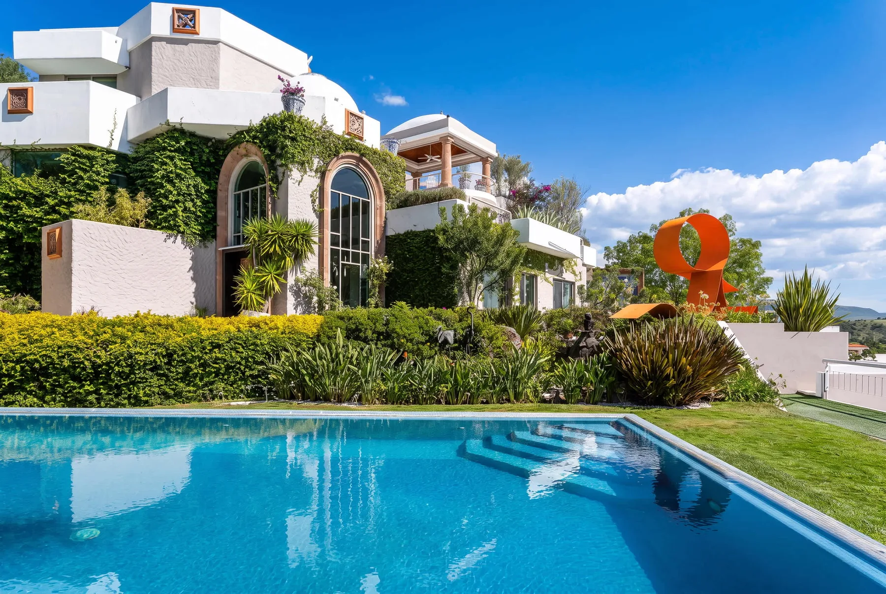 A swimming pool borders a landscaped garden beside a white building under a clear blue sky.