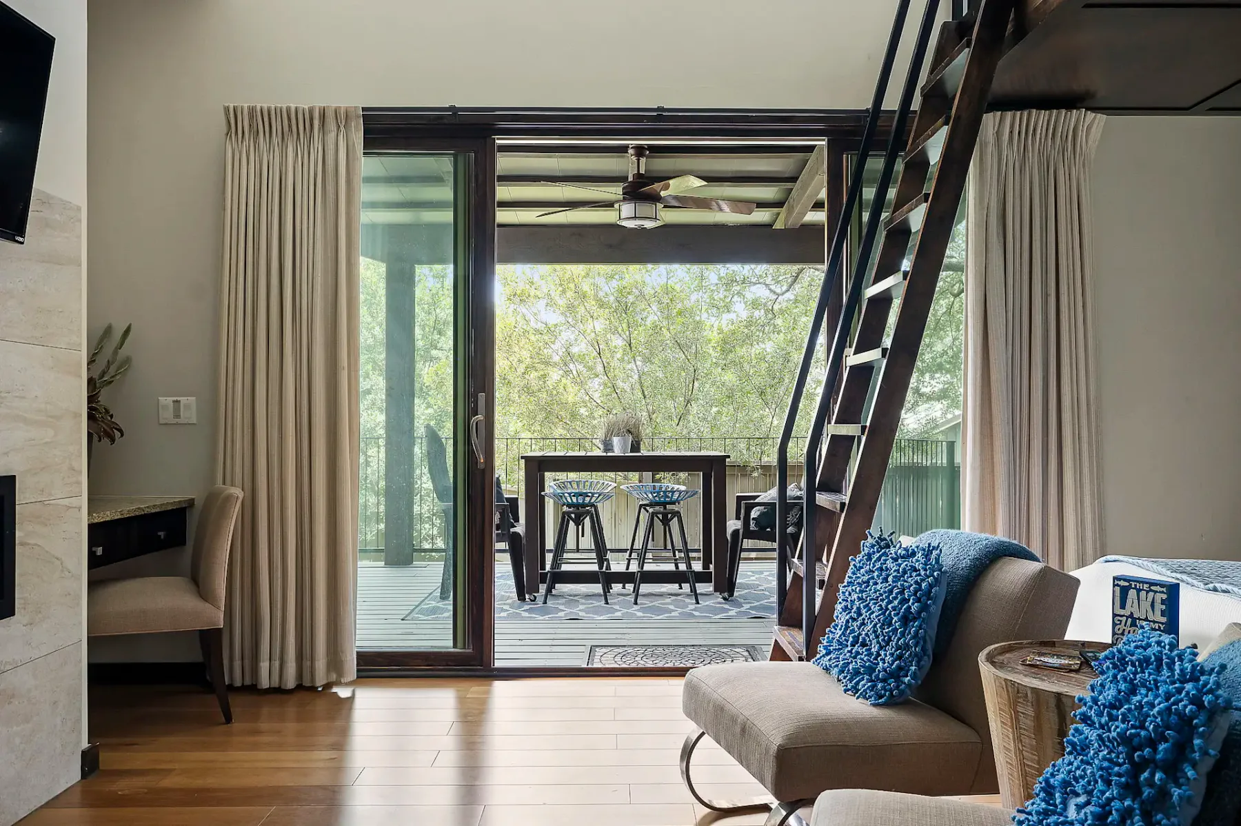 Sala de estar acogedora con cojines azules, una escalera de mano al altillo y puertas corredizas de vidrio que dan a una terraza de madera con asientos al aire libre y vistas al bosque.
