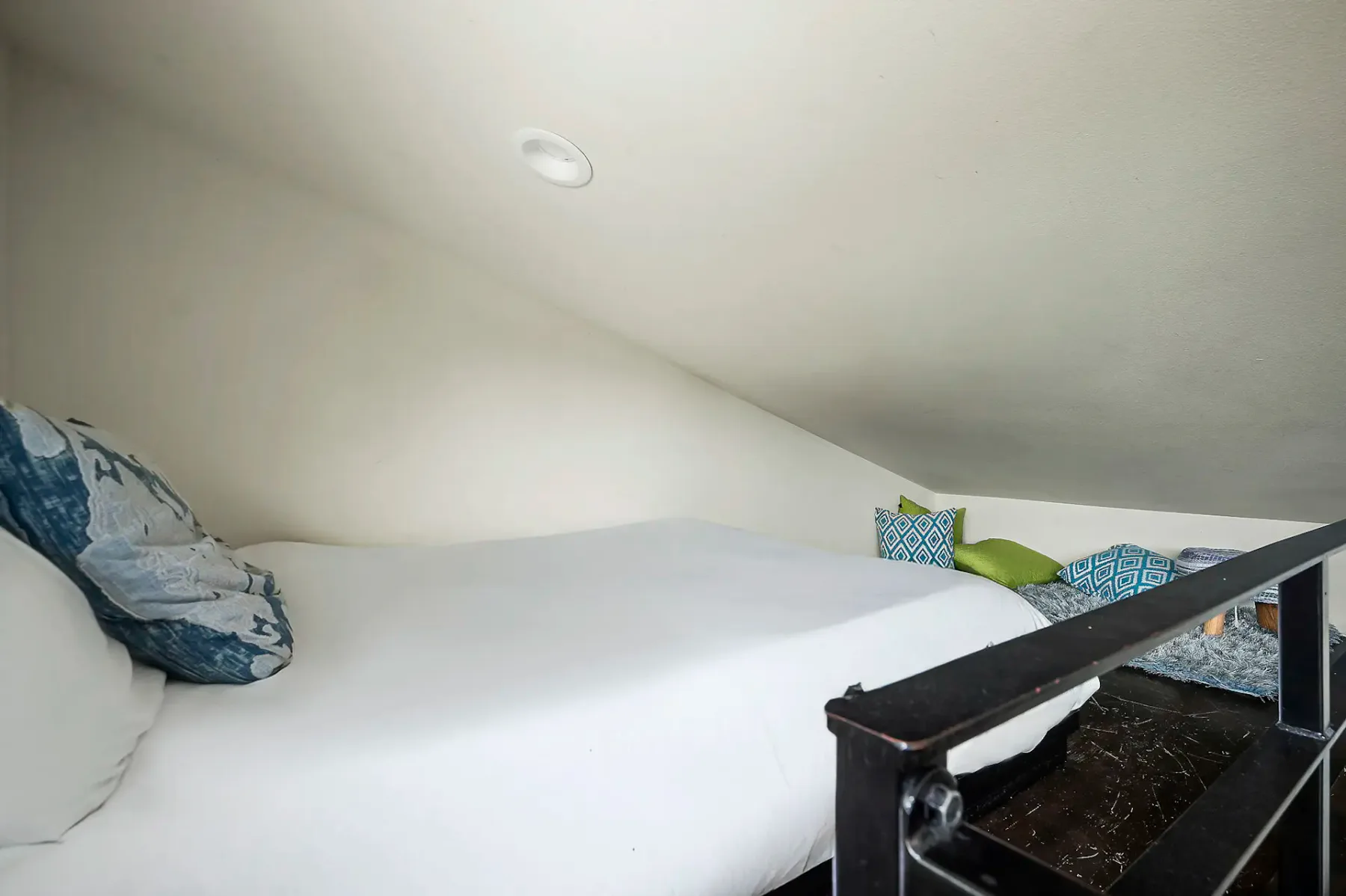 Dormitorio acogedor en un altillo con una cama blanca, cojines con diversos estampados y una barandilla negra bajo un techo inclinado.