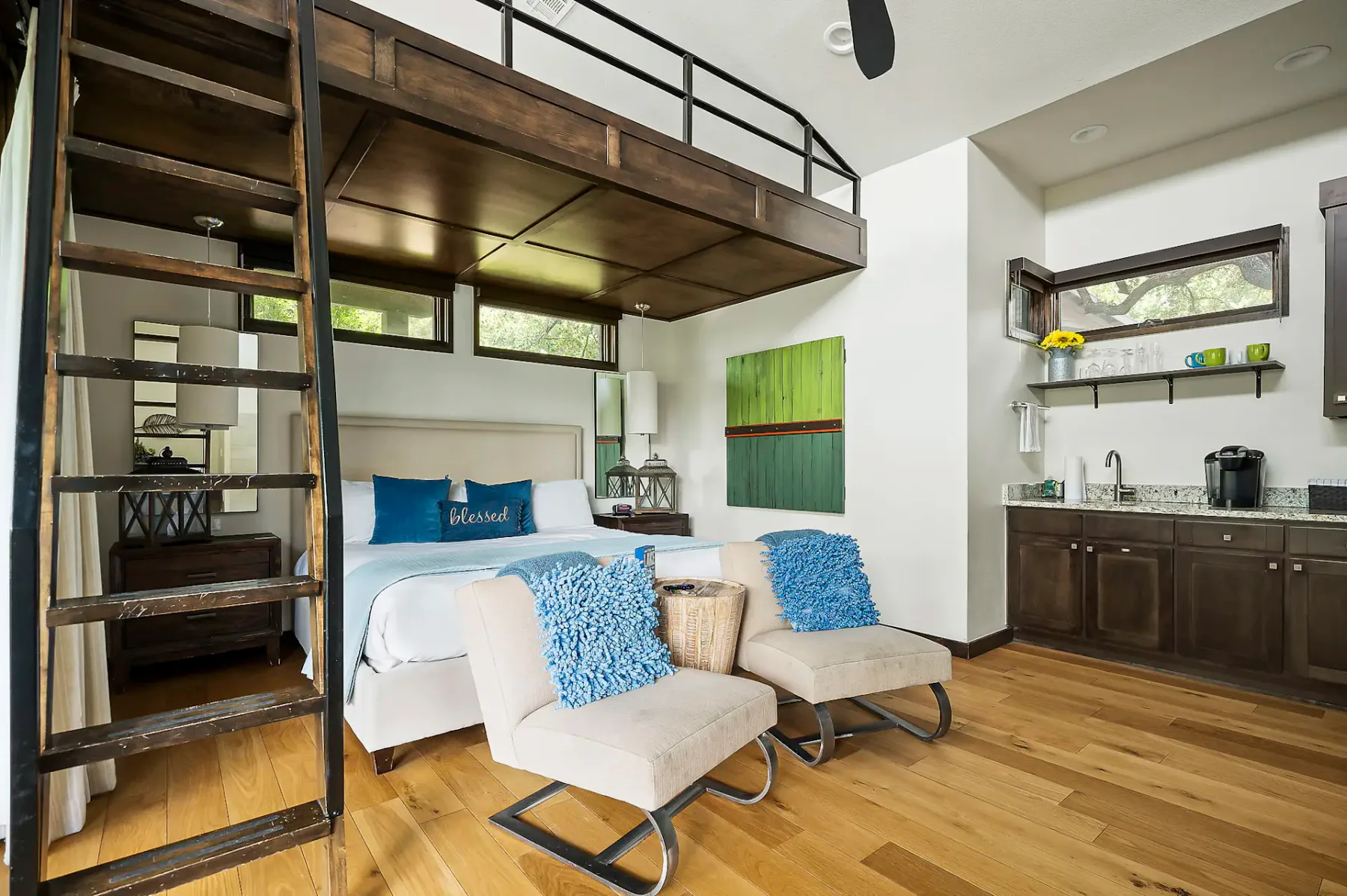 Habitación acogedora con una cama en el altillo, escalera de mano de madera, cama blanca, sillones mullidos con cojines azules, cocina pequeña y decoración moderna sobre un piso de madera clara.