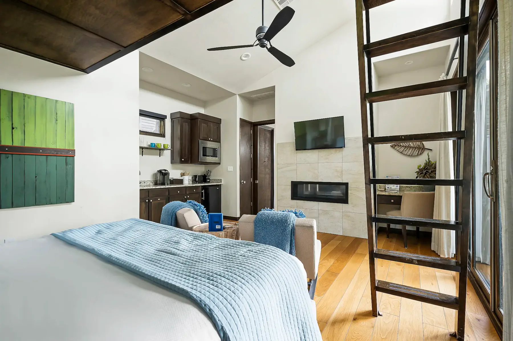 Dormitorio moderno en un altillo con una cama, manta azul, cocina pequeña, escalera de mano, ventilador de techo y luz natural que entra por ventanas grandes. Piso de madera de tonos cálidos en todo el espacio.